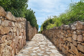 A narrow stone pathway flanked by stone walls and lush greenery, leading to a distant view. - Villa 'Nica' with Private Terrace and Private Garden (Golfo Aranci)