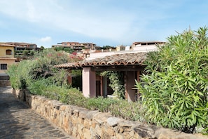 A traditional stone-walled courtyard with a tiled roof structure, surrounded by lush greenery and residential buildings in the background. - Villa 'Nica' with Private Terrace and Private Garden (Golfo Aranci)