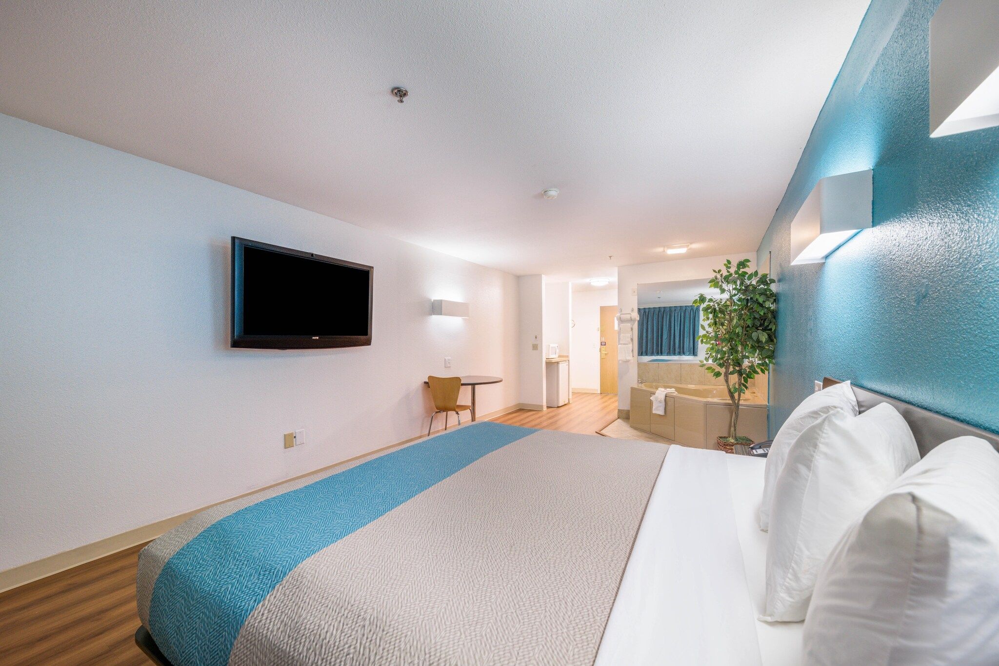 A hotel room with a bed, a television mounted on the wall, a small table with a chair, and a bathroom visible through an open door.