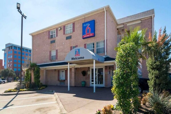 A 6 extended stay hotel with a 'Welcome' sign, a parking lot, and a building with a brick facade. - 1 King Bed | Suite, Non-Smoking, Kitchen (Plano)