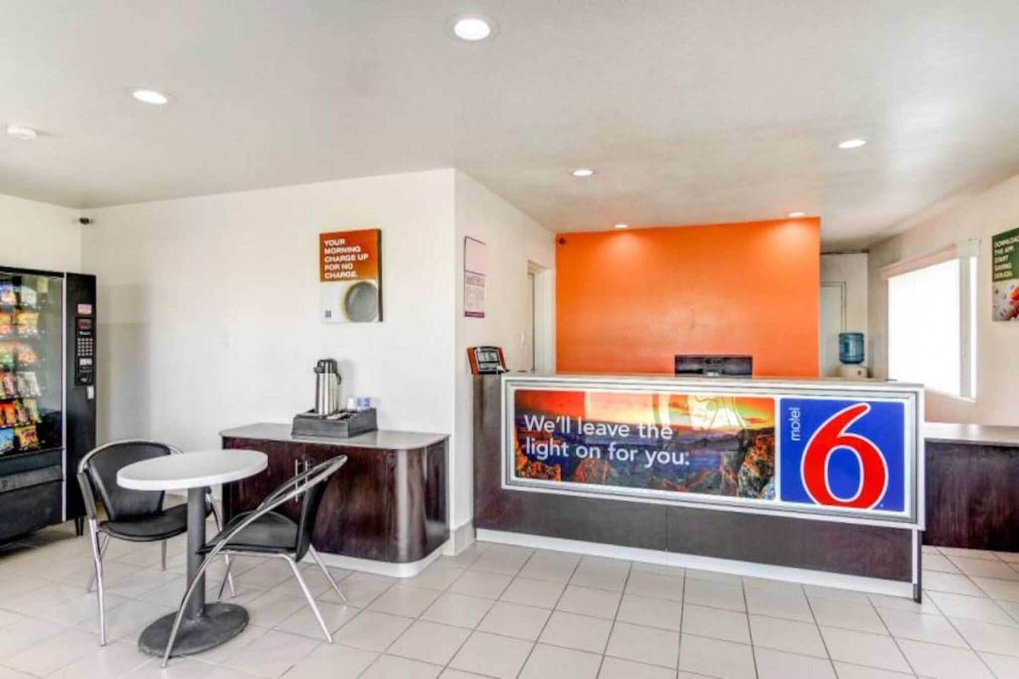 A hotel reception area with a counter, a coffee machine, a refrigerator, and a seating area.