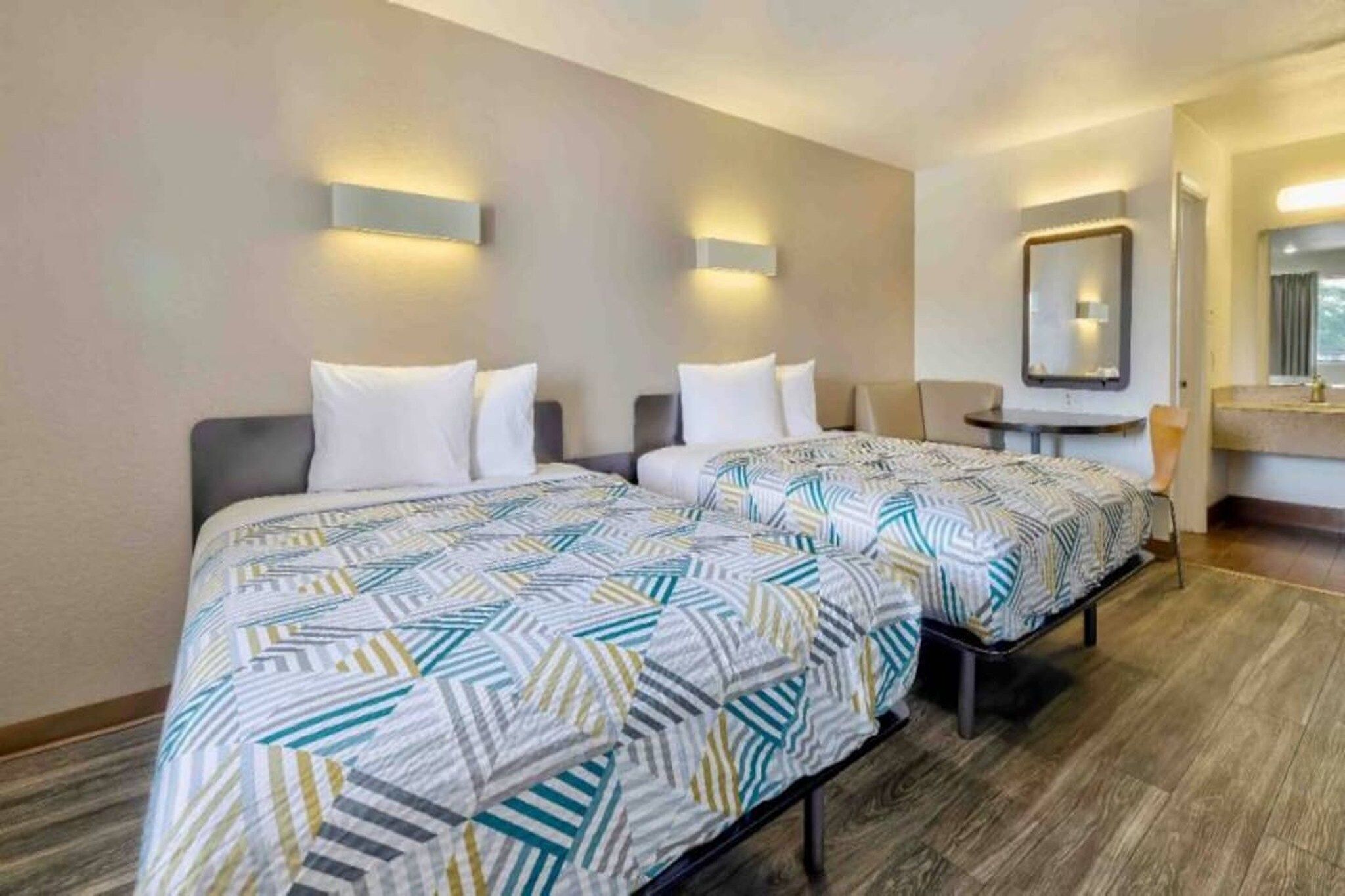 A hotel room with two beds, a small table, a mirror, and a bathroom.