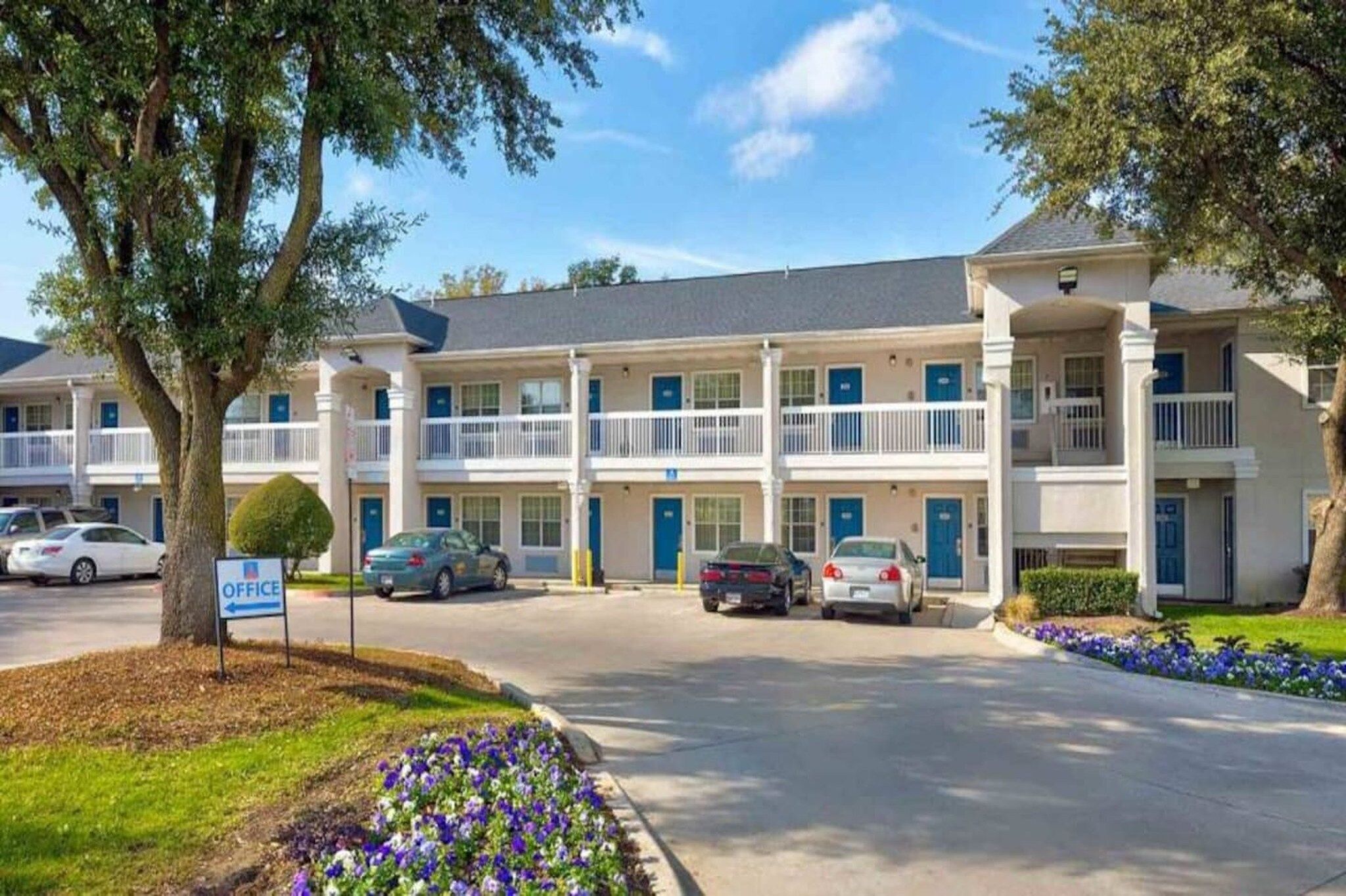 Un motel à plusieurs étages, doté d’un parking, d’une enseigne indiquant la présence de bureaux, et d’une pelouse bien entretenue agrémentée de fleurs.