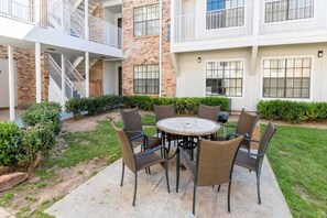 A courtyard with a round table and chairs, surrounded by hedges and a brick building with balconies. - 2 Full Beds |Suite,Nsmk,Kitchen (Arlington)