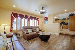 A living room with a brown leather sofa, a round coffee table with a vase of sunflowers, and a view of the kitchen through a window with red curtains. - Patio & Grill! Spacious Las Vegas Oasis (Las Vegas)