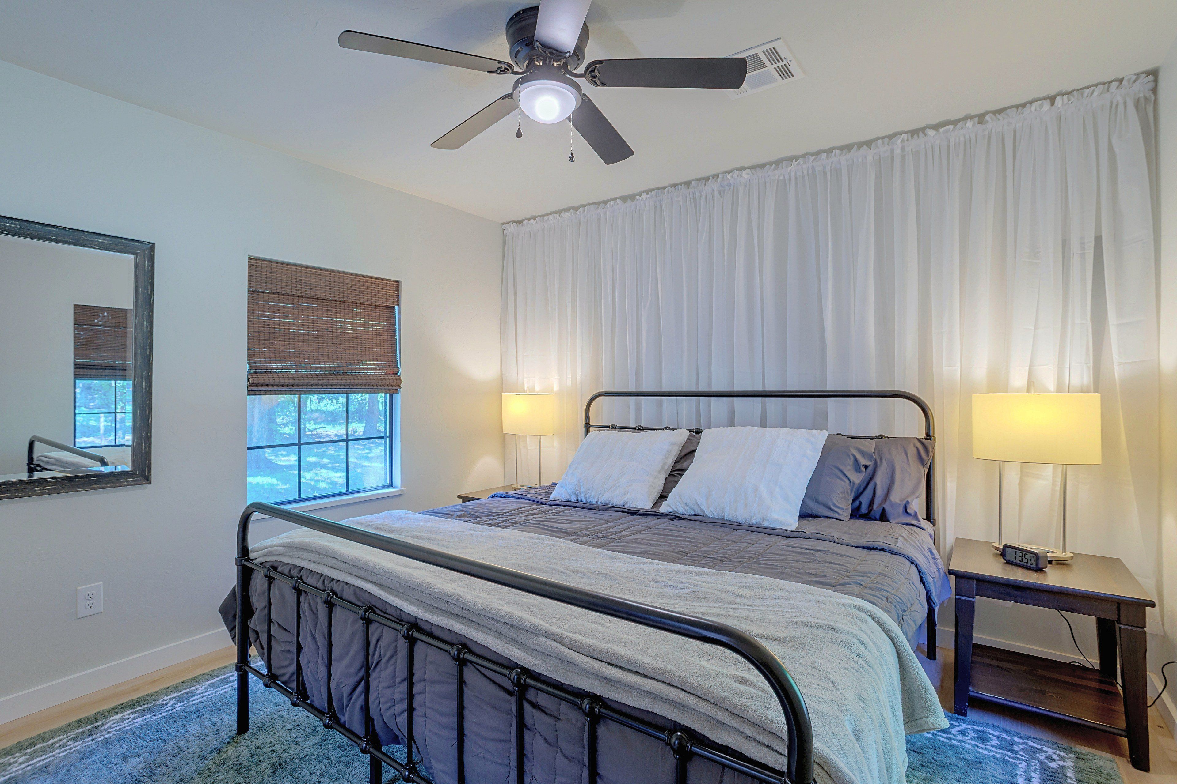 A bedroom with a bed, a ceiling fan, a window with blinds, a bedside table with a lamp, and a wall-mounted mirror.
