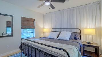 A bedroom with a bed, a ceiling fan, a window with blinds, a bedside table with a lamp, and a wall-mounted mirror.