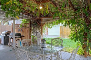 Eine Terrasse im Freien mit einem Glastisch und weißen Stühlen, umgeben von Grünpflanzen und einem Holzzaun.