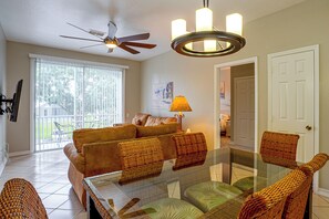 A living room with a glass-top coffee table, wicker chairs, a brown sofa, a ceiling fan, and a door leading to another room. - Resort Perks! Family Condo 4 Mi to Disney World! (Kissimmee)