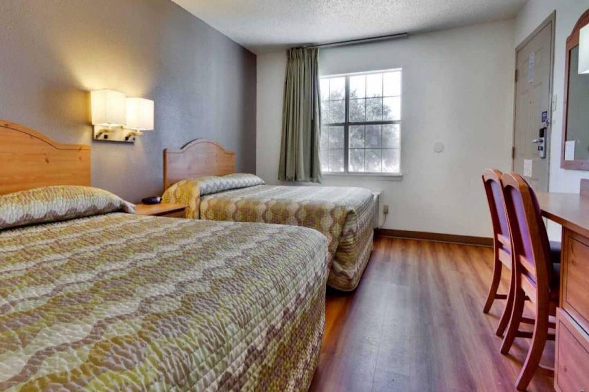 A hotel room with two beds, a desk, a chair, a window with curtains, and a lamp.