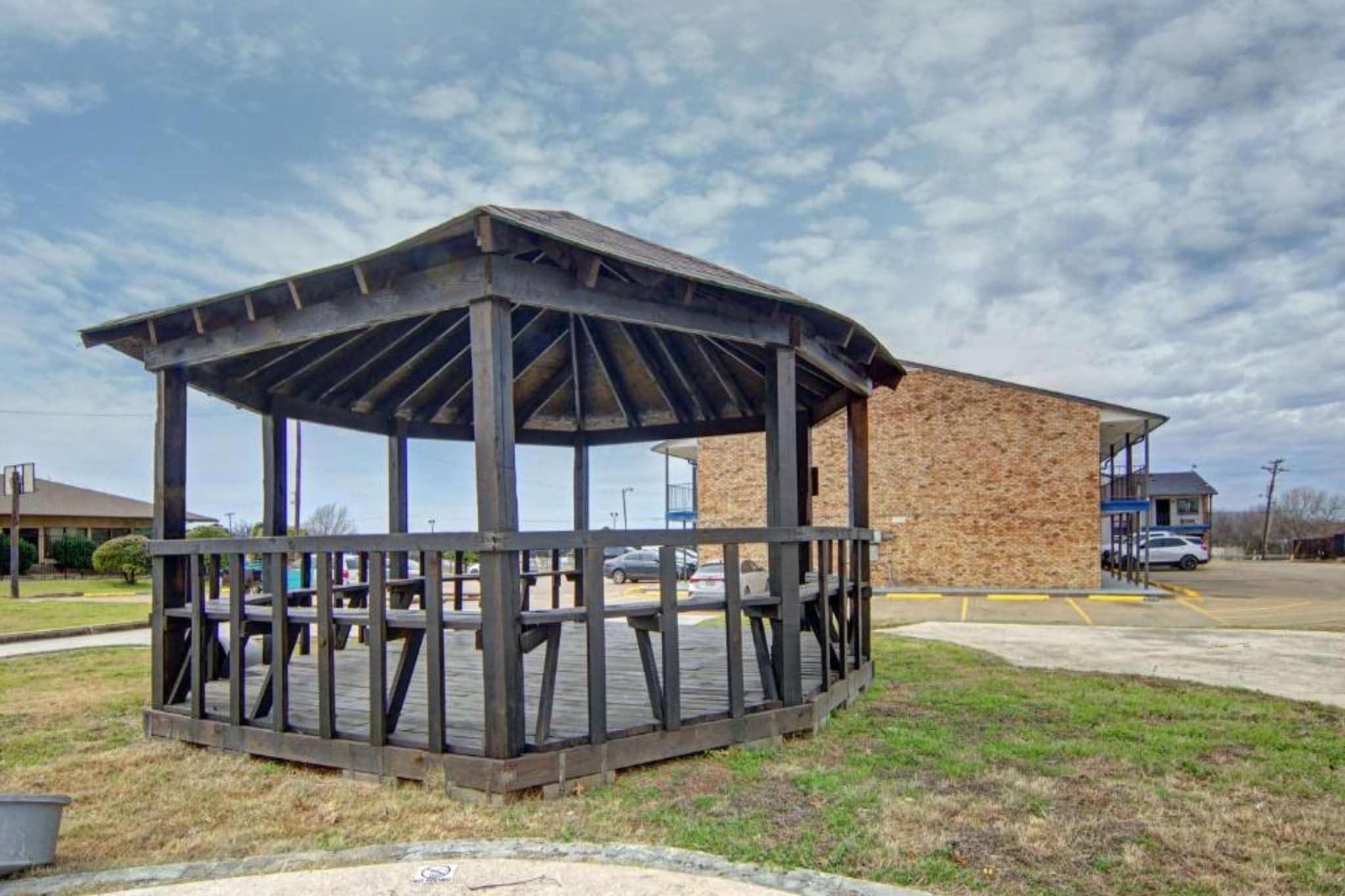 A gazebo with a wooden deck, a brick building in the background, and a parking lot.
