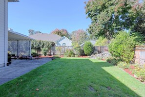 A residential backyard with a lawn, a covered patio area, and various plants and trees. - Lovely, Spacious Gem: Walk to Autzen & River Trail (Springfield)