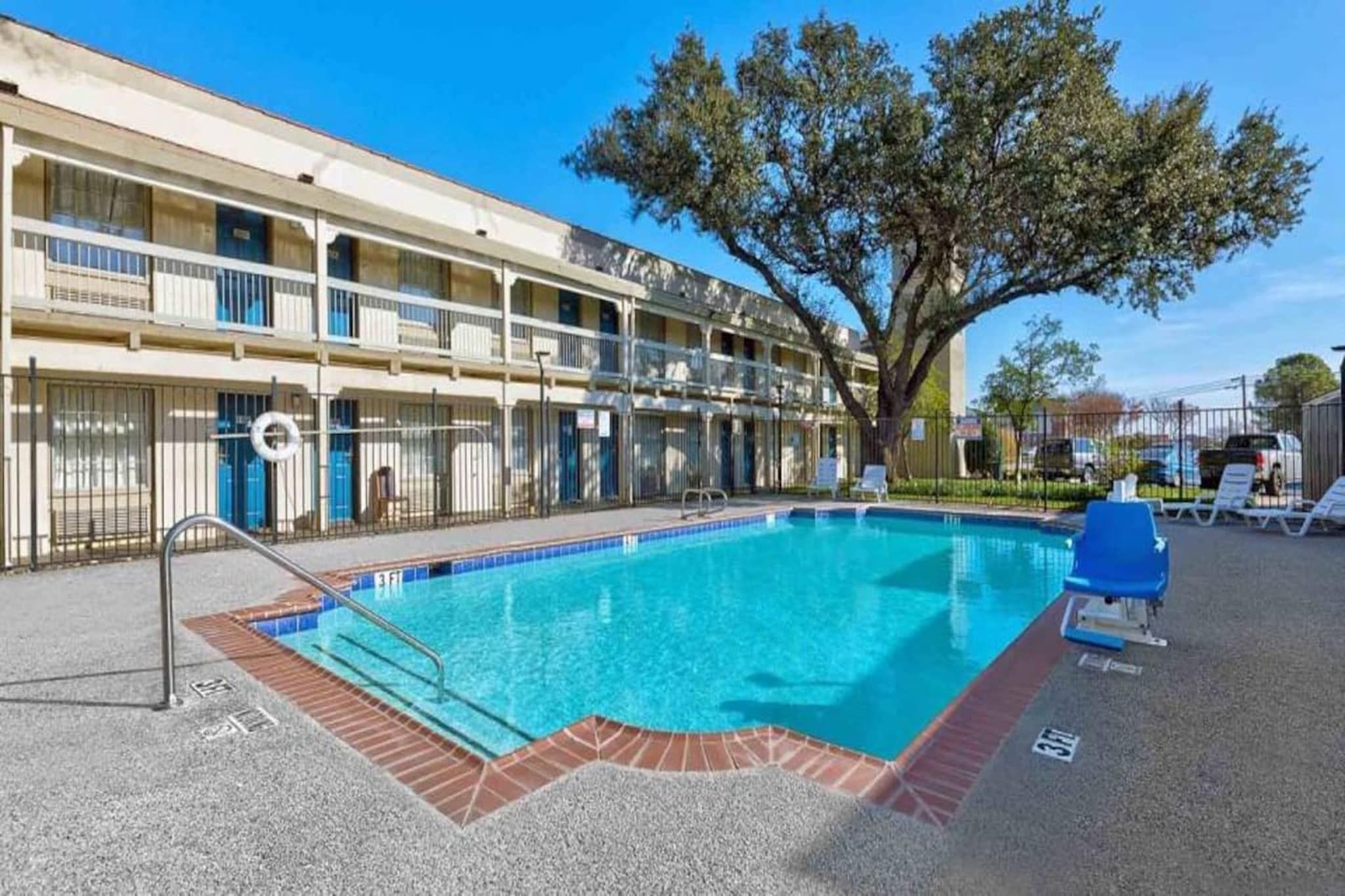 A swimming pool with a clear blue water, surrounded by a fenced area, and a multi-story building with balconies in the background.