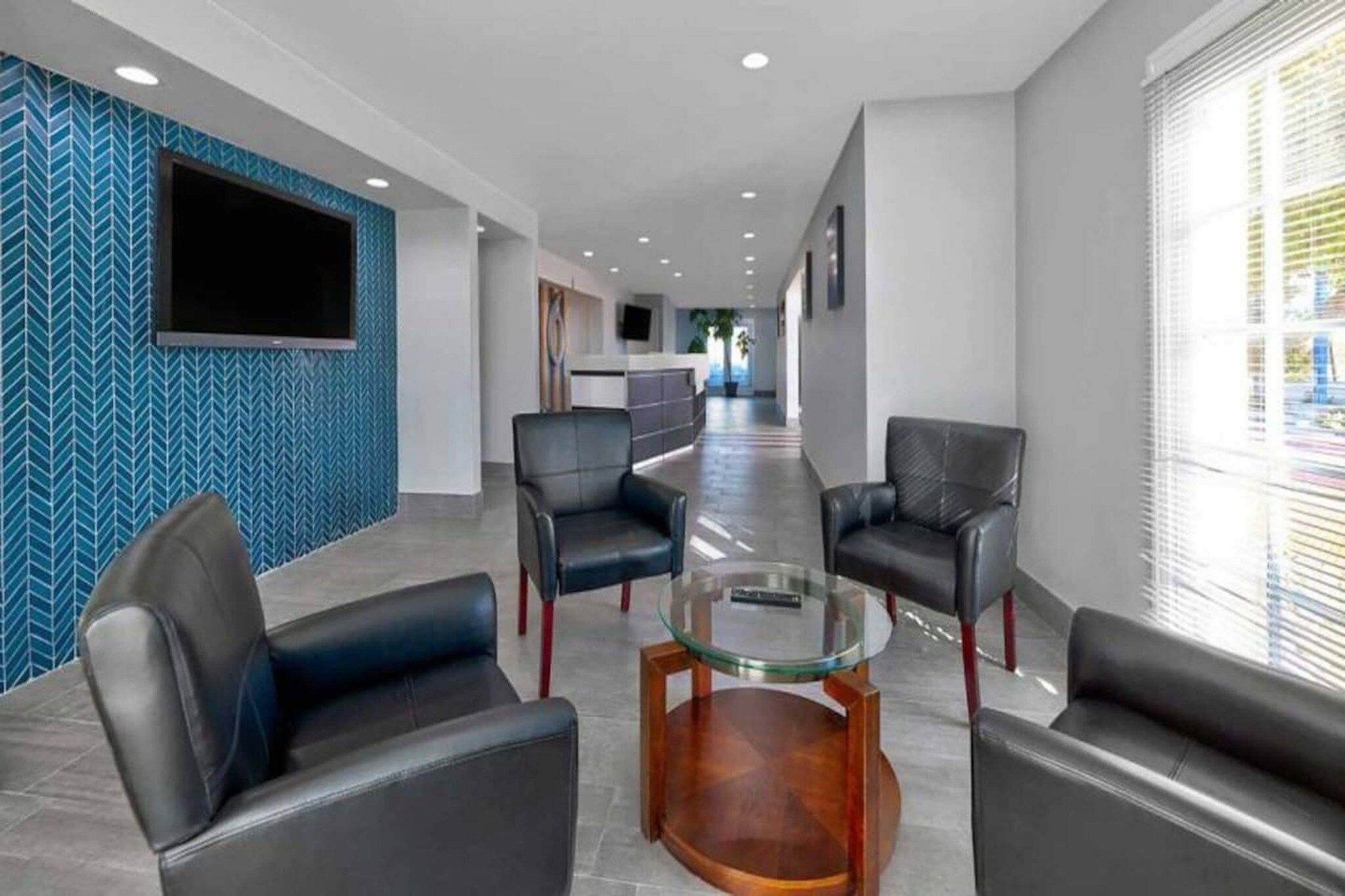 A modern lobby with a central glass table surrounded by leather chairs, a reception desk in the background, and a patterned wall on one side.