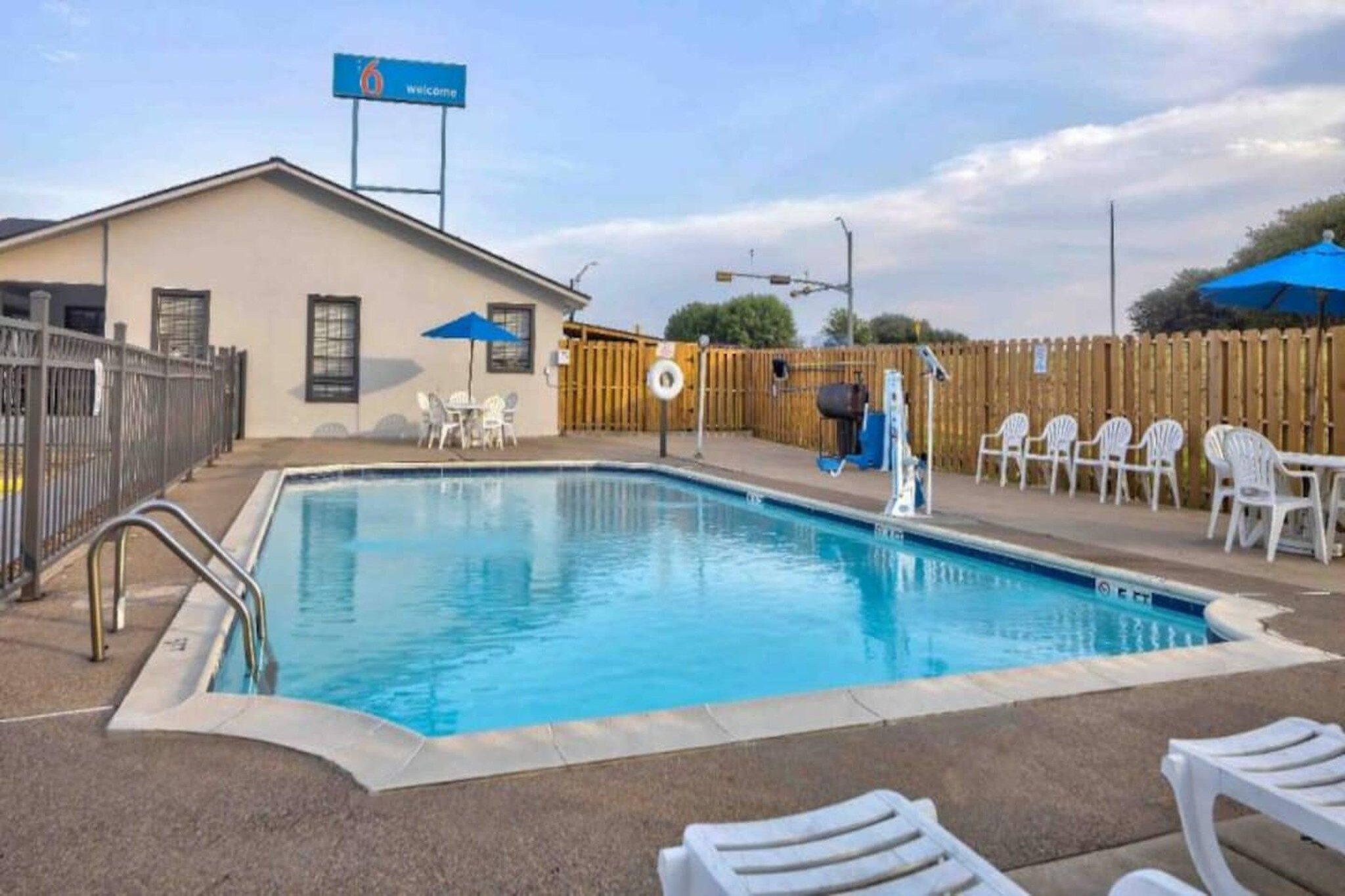 Une piscine entourée d’une clôture en bois, un bâtiment avec une pancarte « Bienvenue », et des sièges à l’extérieur.