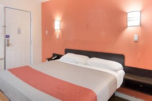 A hotel room with a bed, a nightstand, a door, and two wall-mounted lights. - 1 King Bed | Smoking, Micfridge (Fort Worth)