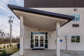 Entrada de un edificio moderno con un vestíbulo cubierto, puerta de cristal y señal de accesibilidad para personas con discapacidad.