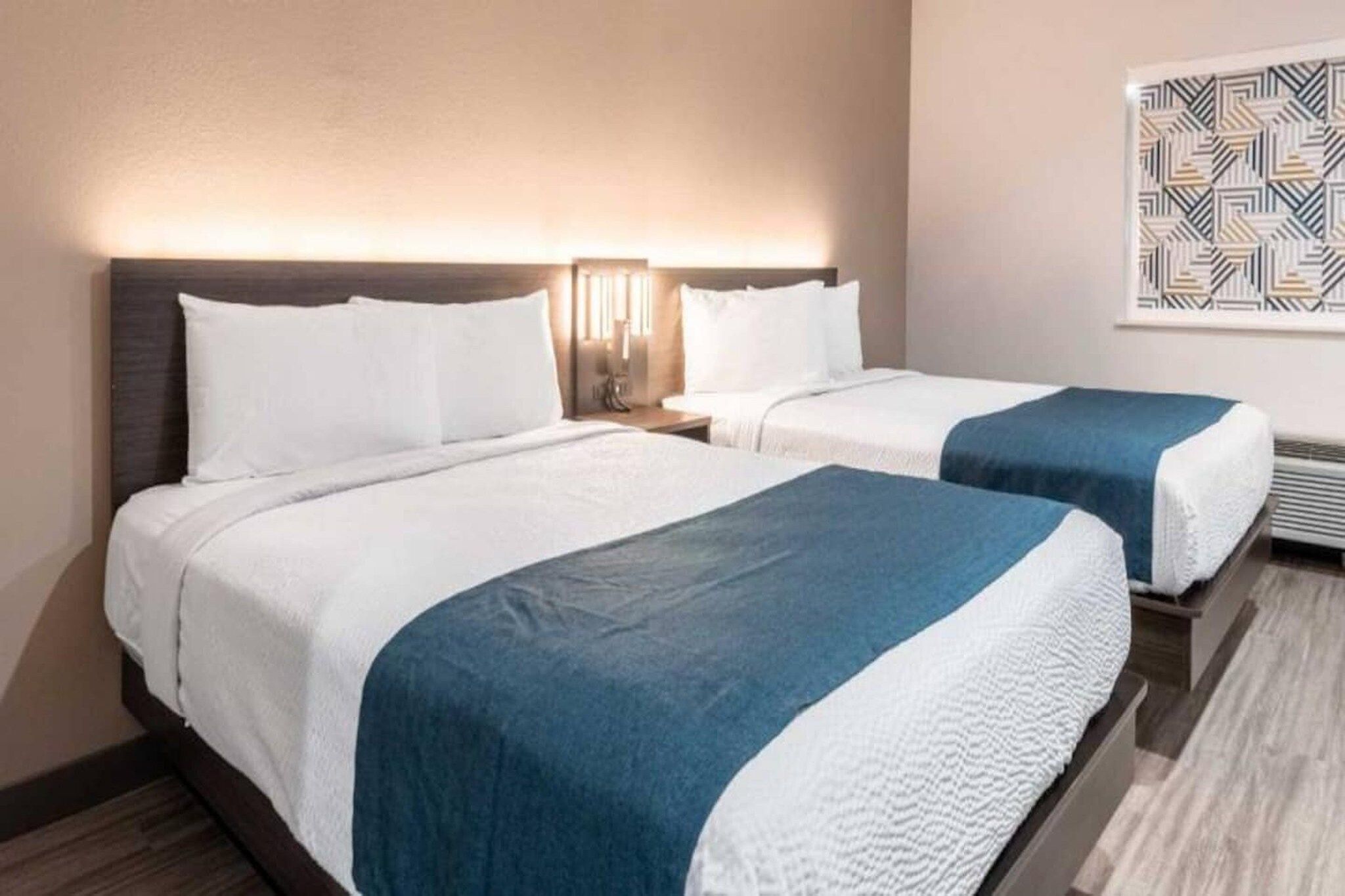 Quarto de hotel com duas camas, cada uma com roupa de cama branca e detalhes azuis. Há uma mesa de cabeceira com abajur entre as camas.
