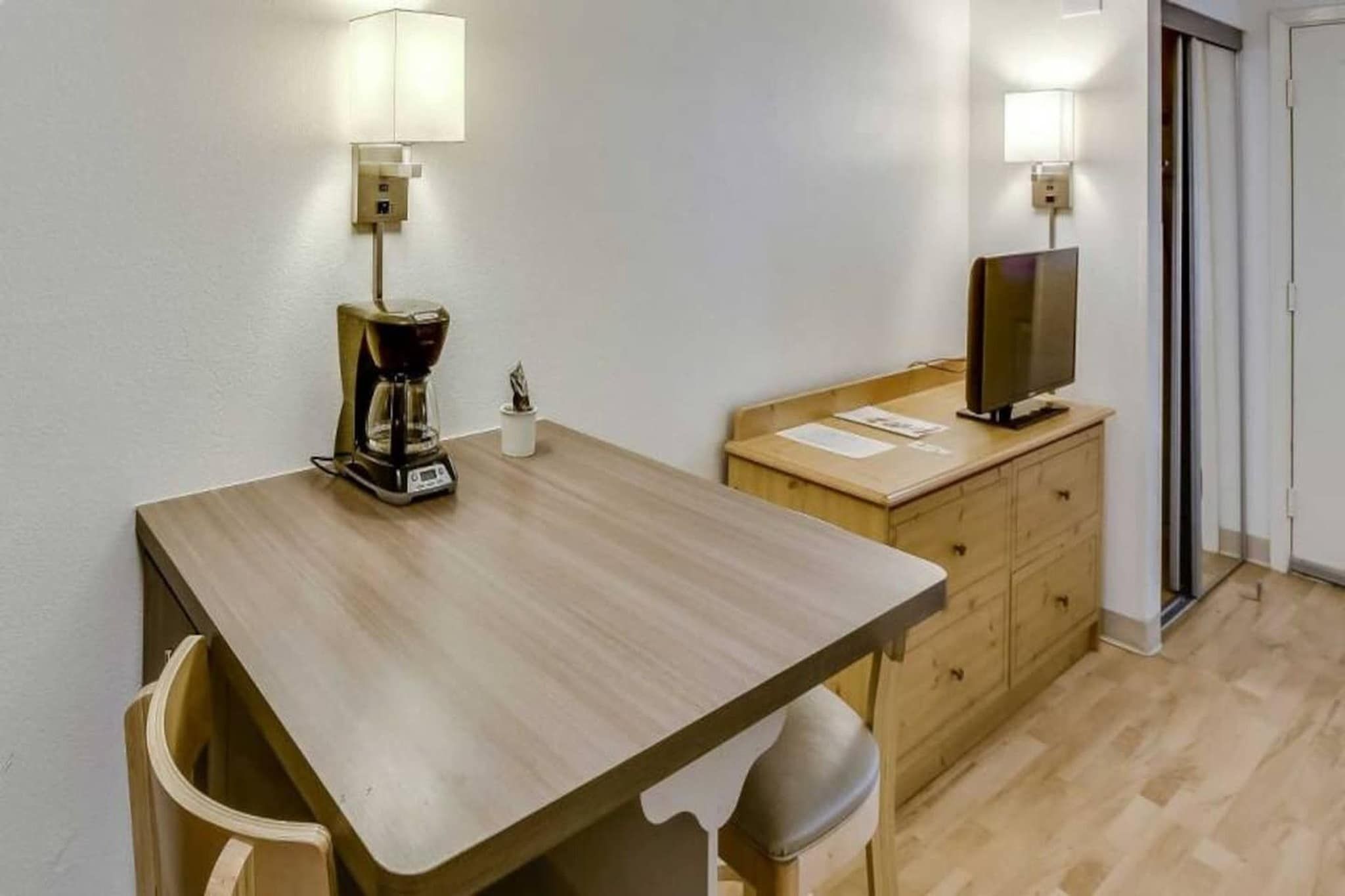 A small, simple room with a wooden table, a coffee maker, a television on a wooden cabinet, and a door.