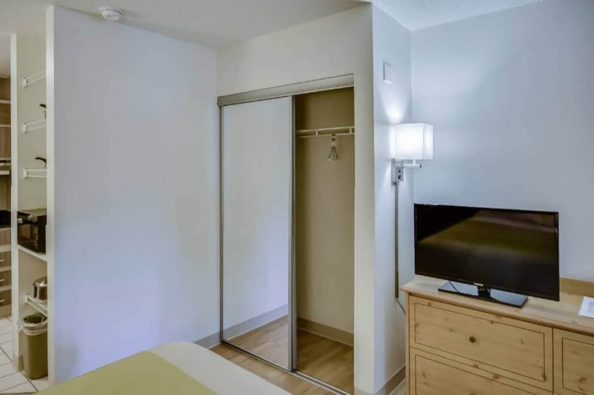 Une chambre à coucher avec un lit, une armoire, un téléviseur posé sur une commode en bois et un miroir accroché au mur.