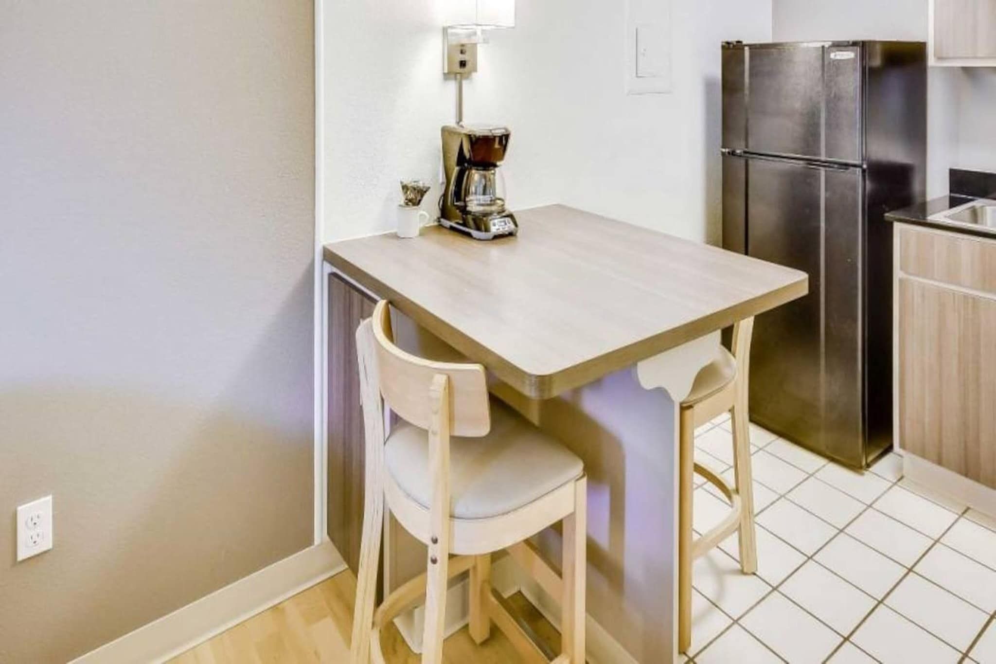 A small kitchen with a wooden table, two chairs, a coffee maker, and a stainless steel refrigerator.