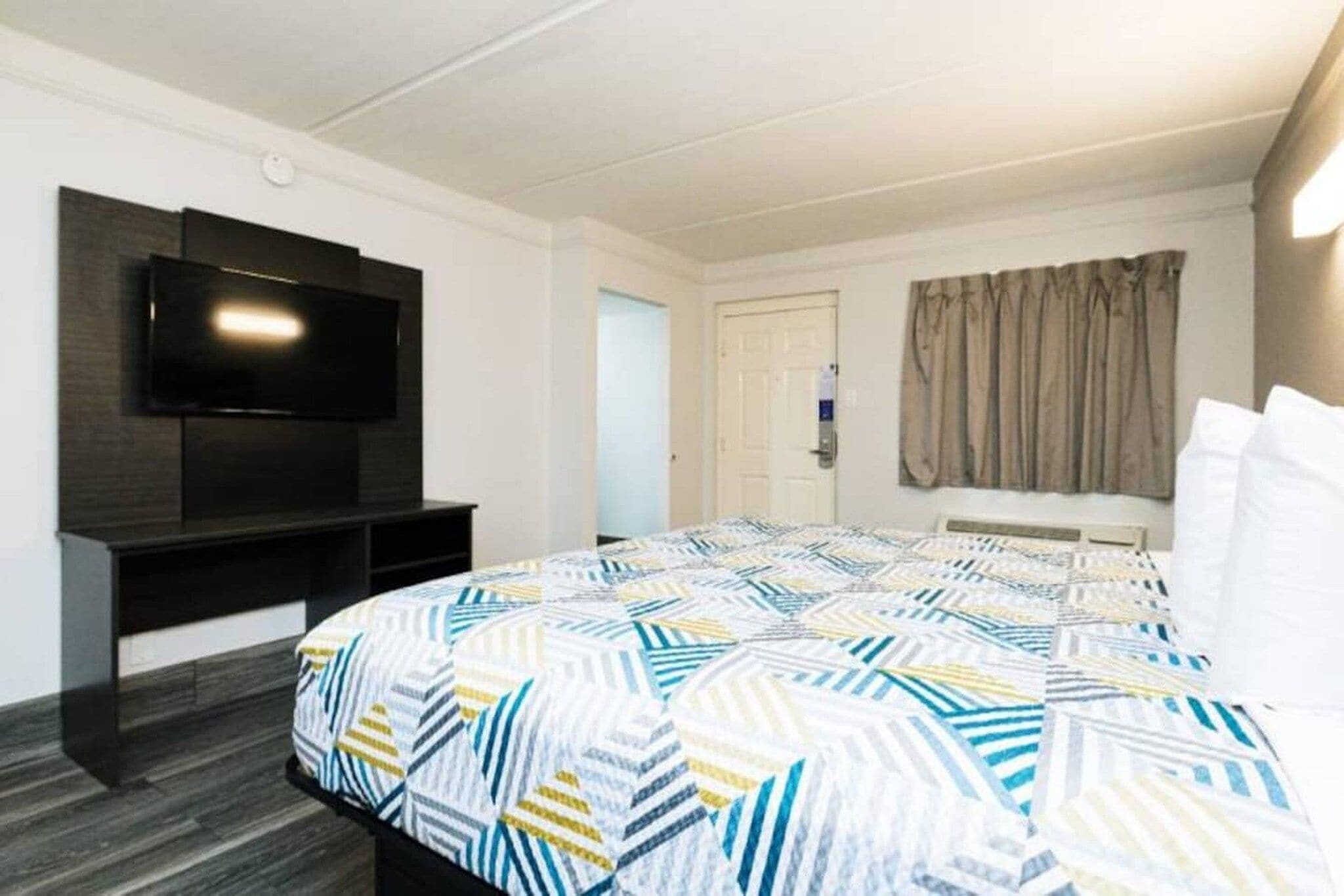 A hotel room with a bed, a flat-screen TV mounted on the wall, a door, and a window with curtains.