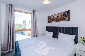 Une chambre d’hôtel avec un grand lit, une tête de lit, une fenêtre avec des rideaux, un tableau au mur et un vase de fleurs sur la table de chevet.