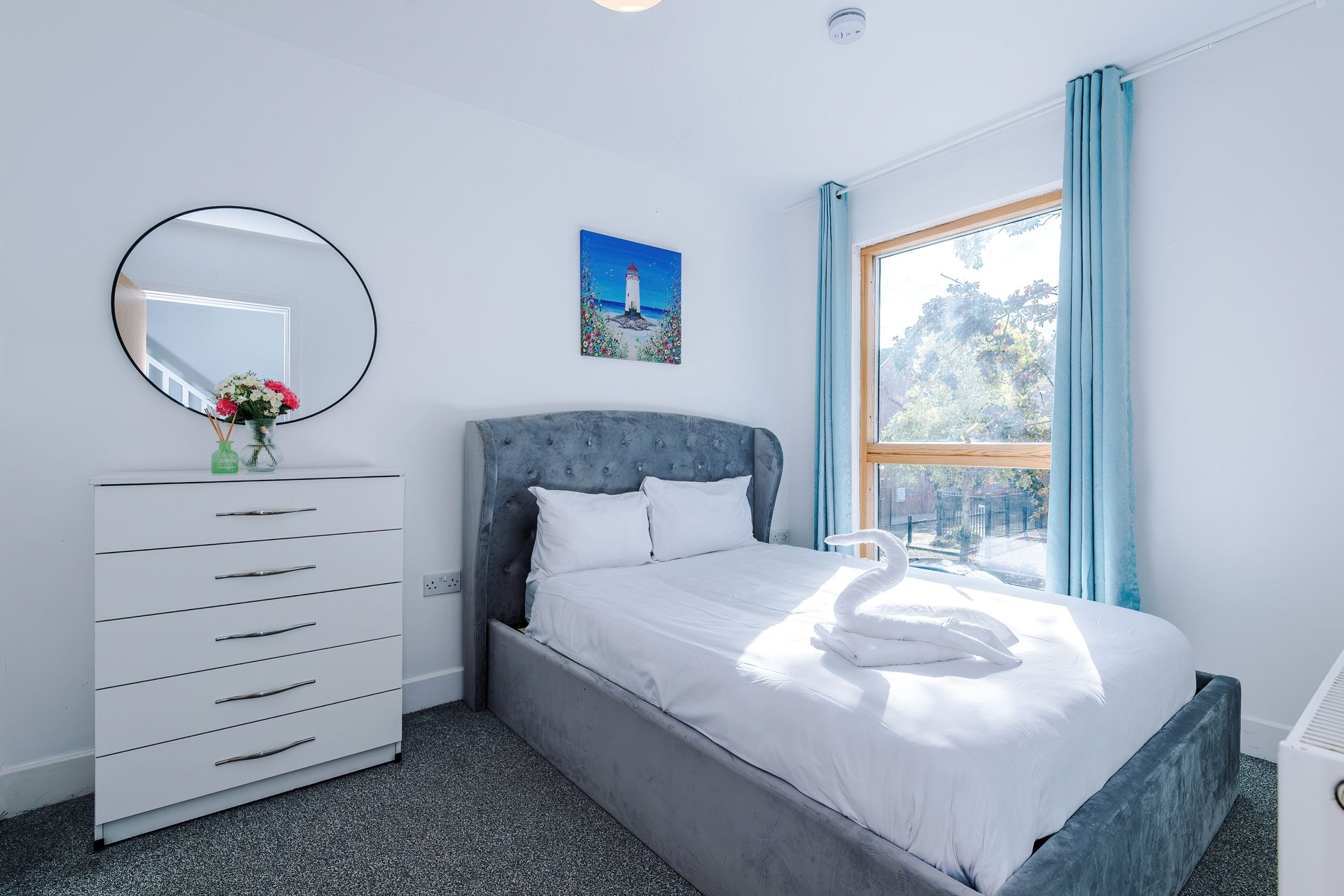 A bedroom with a bed, a dresser, a round mirror, a painting, and a window with curtains.