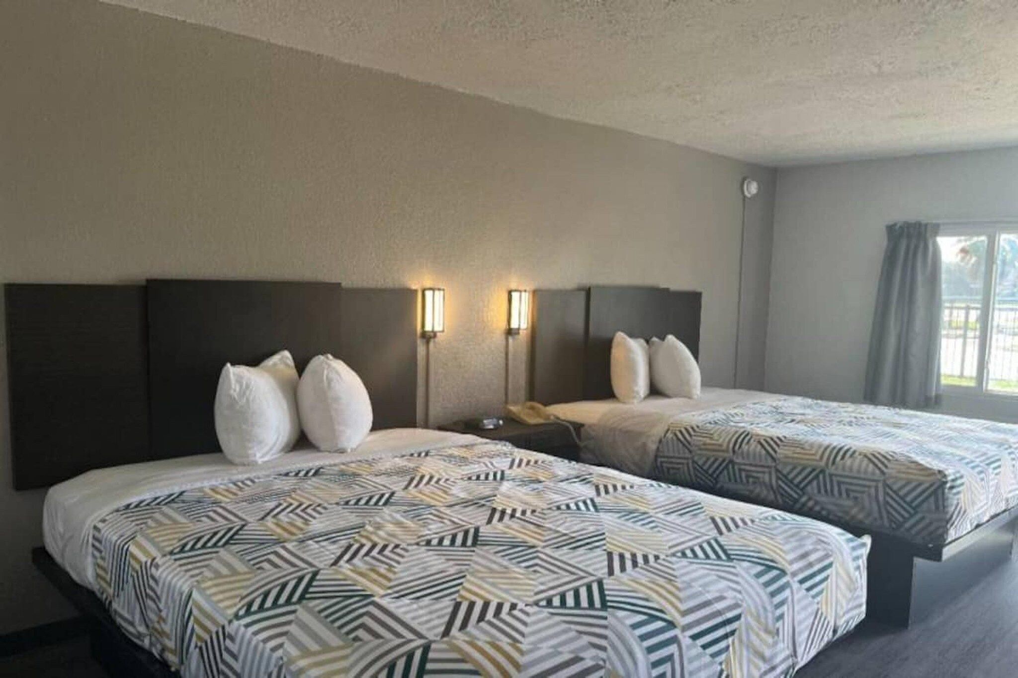 A hotel room with two beds, each with patterned bedding and headboards, a nightstand, and wall-mounted reading lamps.