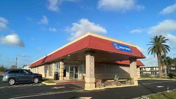 A building with a red roof and a 'NOW OPEN' sign, a parking lot, and a palm tree in the background.