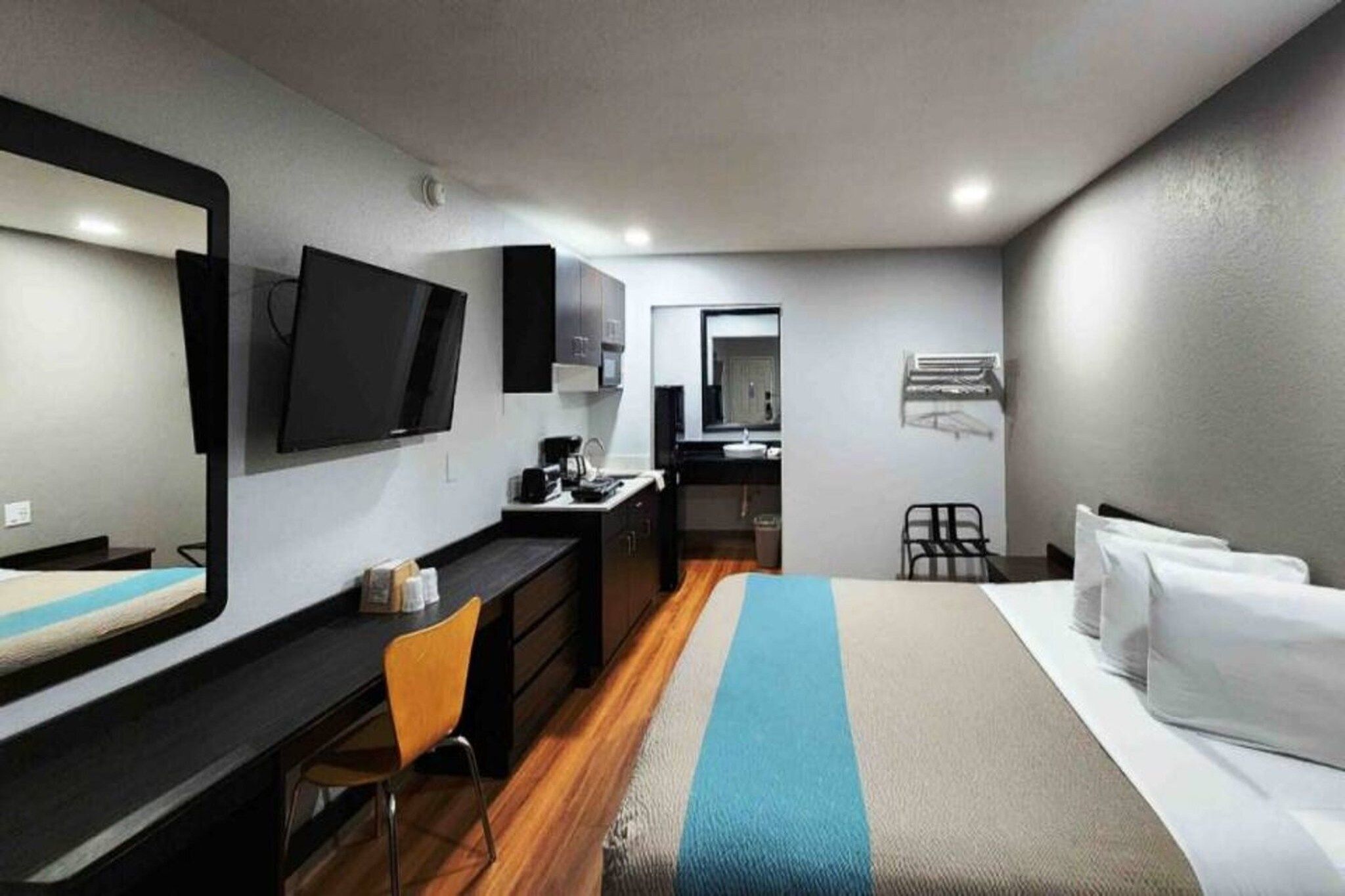 Ett hotellrum med en säng, ett skrivbord, en stol, en tv, en spegel och ett litet kök.