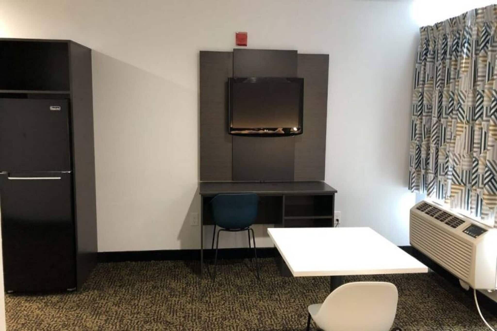 A compact room with a desk, a chair, a small table, a refrigerator, and an air conditioning unit.