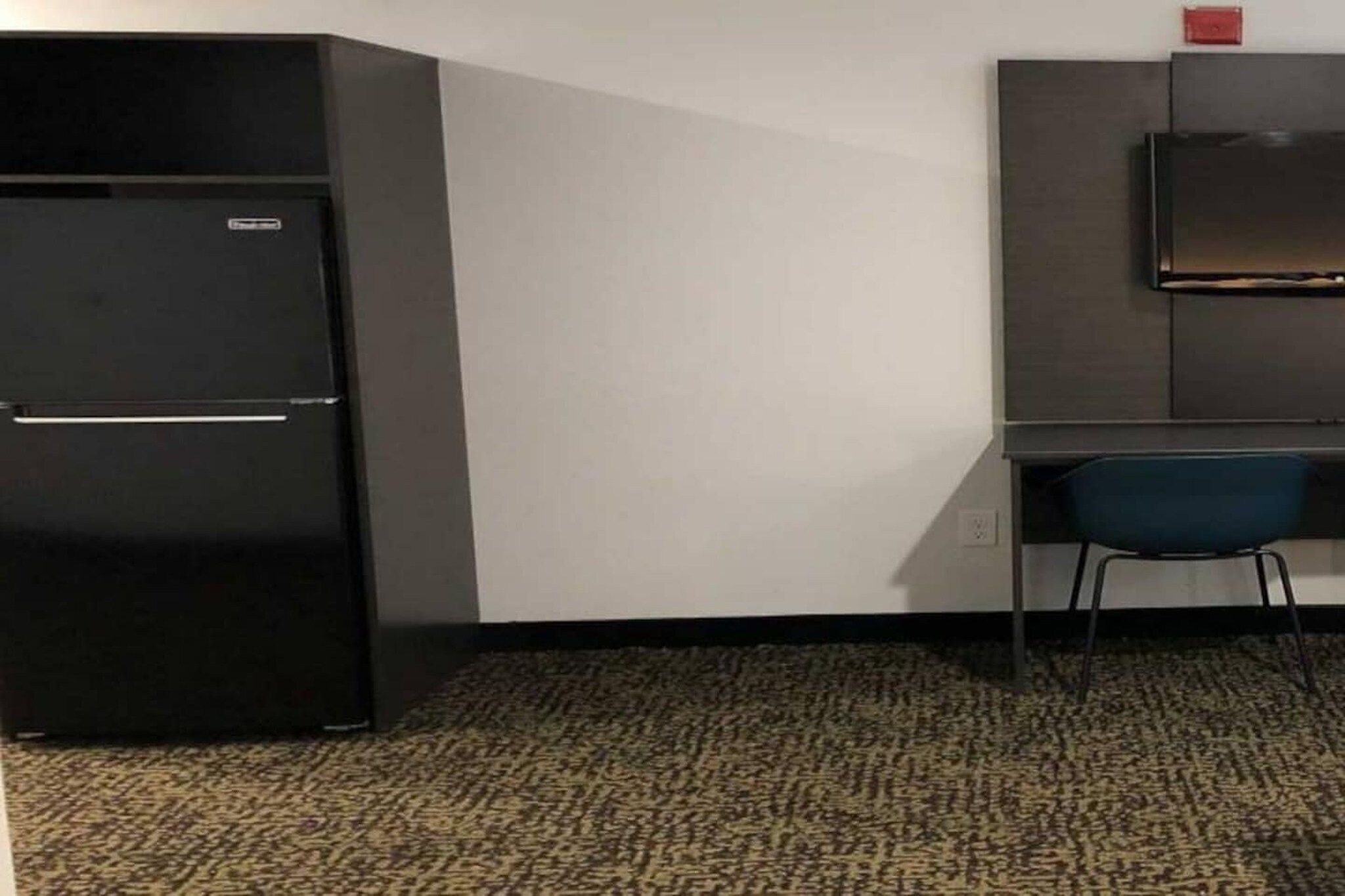 A compact room with a black refrigerator, a desk with a chair, and a flat-screen TV mounted on the wall.