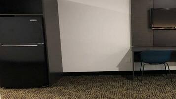 A compact room with a black refrigerator, a desk with a chair, and a flat-screen TV mounted on the wall.