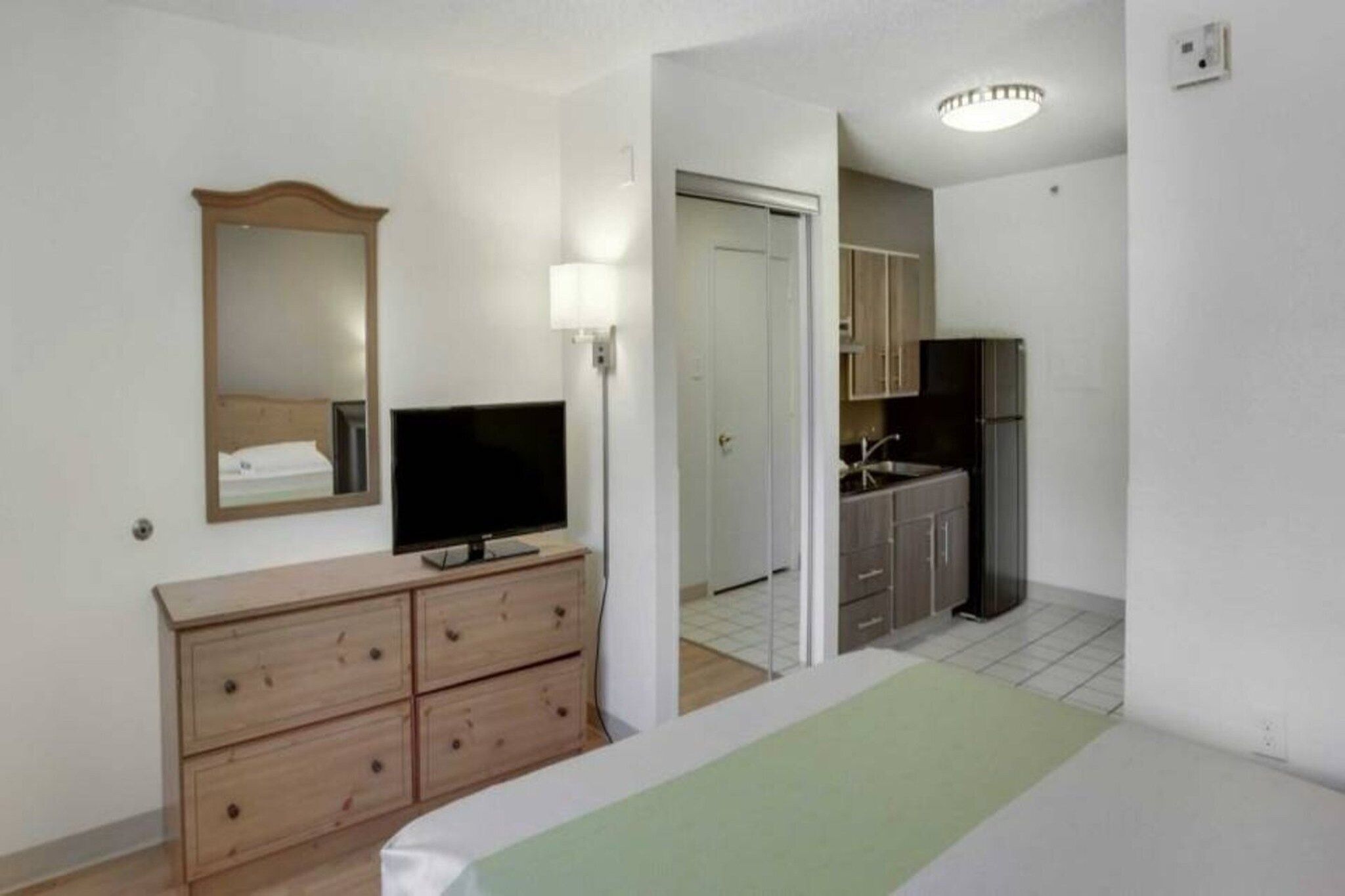 A hotel room with a bed, a dresser, a television, a mirror, a door, and a kitchenette.