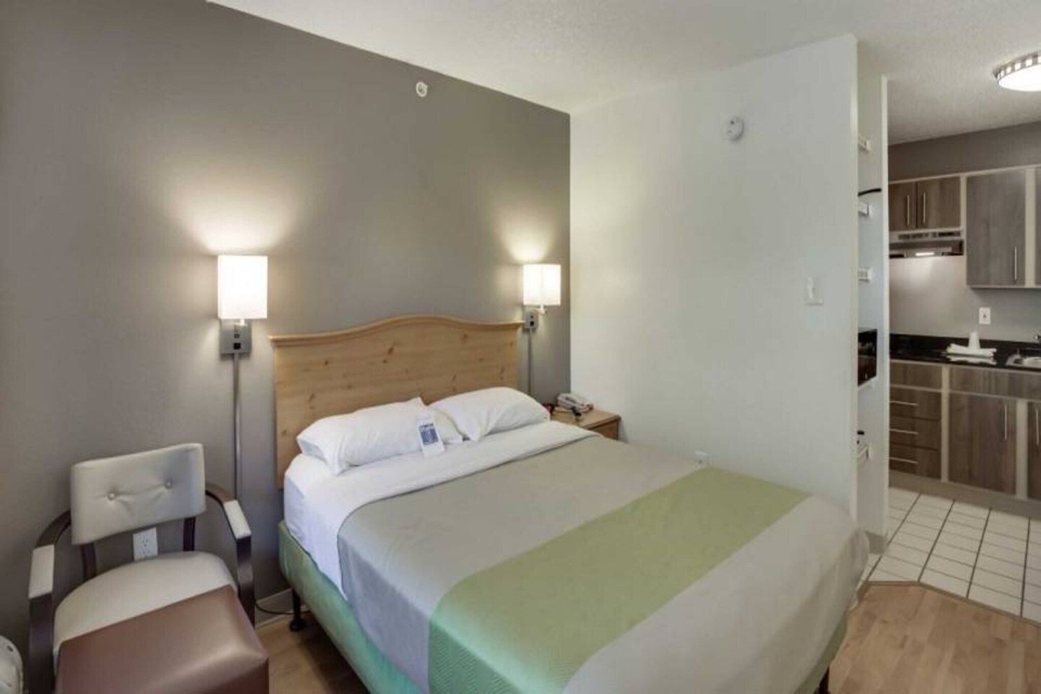 A hotel room with a bed, bedside lamps, a chair, and a kitchenette.