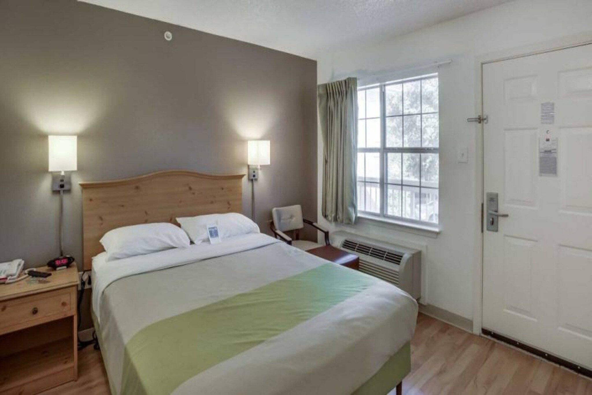 A hotel room with a bed, bedside lamps, a nightstand, a window with curtains, and a door.