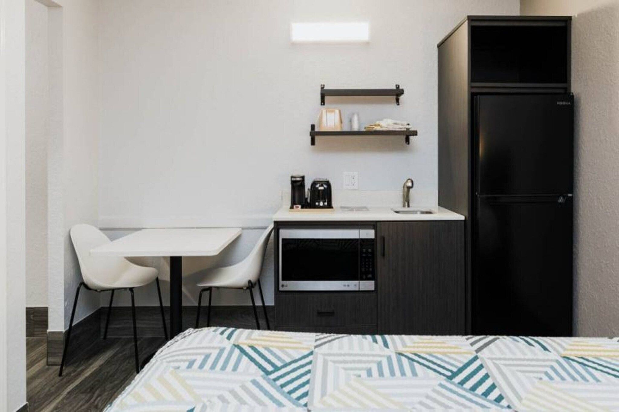 A compact kitchenette with a microwave, coffee maker, and a small refrigerator. There is a white table with two chairs and a bed with a patterned bedspread.
