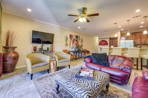 A living room with a ceiling fan, a flat-screen TV, wicker furniture, a red leather sofa, and a zebra-patterned ottoman. - Pool & River Access! Luxury Hill Country Haven (New Braunfels)