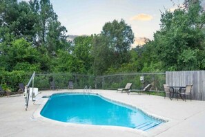 Une piscine rectangulaire avec un escalier en métal, entourée d’une clôture. On y trouve des chaises longues et une table près de la piscine.