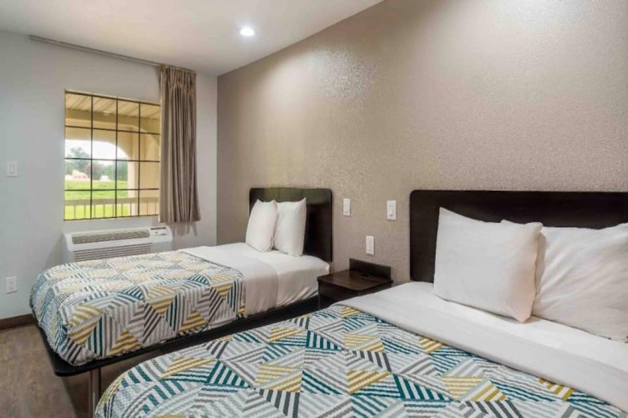 A hotel room with two beds, a window with curtains, an air conditioning unit, and a view of the outdoors.