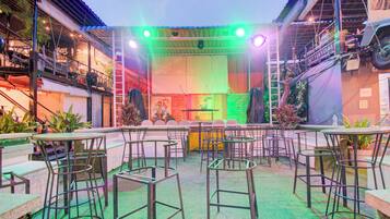 An outdoor seating area with tables and chairs, a stage with colorful lighting, and a brick wall backdrop.