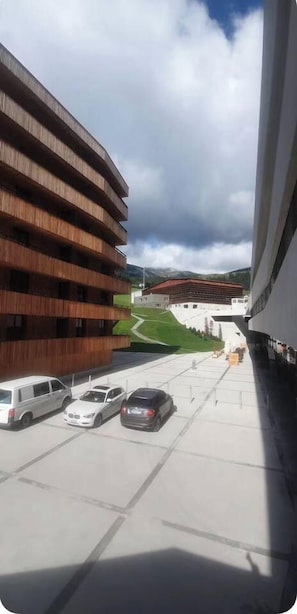 A modern building with a curved facade, a parking area with several cars, and a view of a distant building with a red roof. - Central 2.5-Room Apartment in Flims - Ski-in/ski-out (Flims)