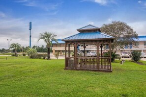 Un kiosque doté d’un toit en métal, entouré d’une pelouse bien entretenue, avec un bâtiment et des voitures garées en arrière-plan.