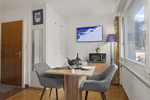 A compact room with a wooden dining table, two chairs, a television mounted on the wall, a microwave, and a window with curtains. - Central studio apartment, close to the ski lifts (Crans-Montana)
