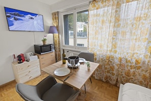 A compact room with a wooden dining table, a single chair, a television mounted on the wall, and a window with curtains. - Central studio apartment, close to the ski lifts (Crans-Montana)