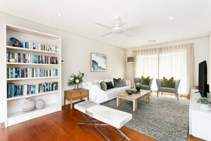 A modern living room with a bookshelf, a white sofa, a coffee table, and a television. - EDBAY - Edwards Beach Retreat | Steps To Balmoral (Mosman)