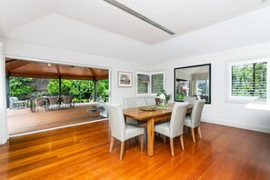 A spacious dining area with a wooden table and chairs, large windows with blinds, and a view of a lush garden. - EDBAY - Edwards Beach Retreat | Steps To Balmoral (Mosman)
