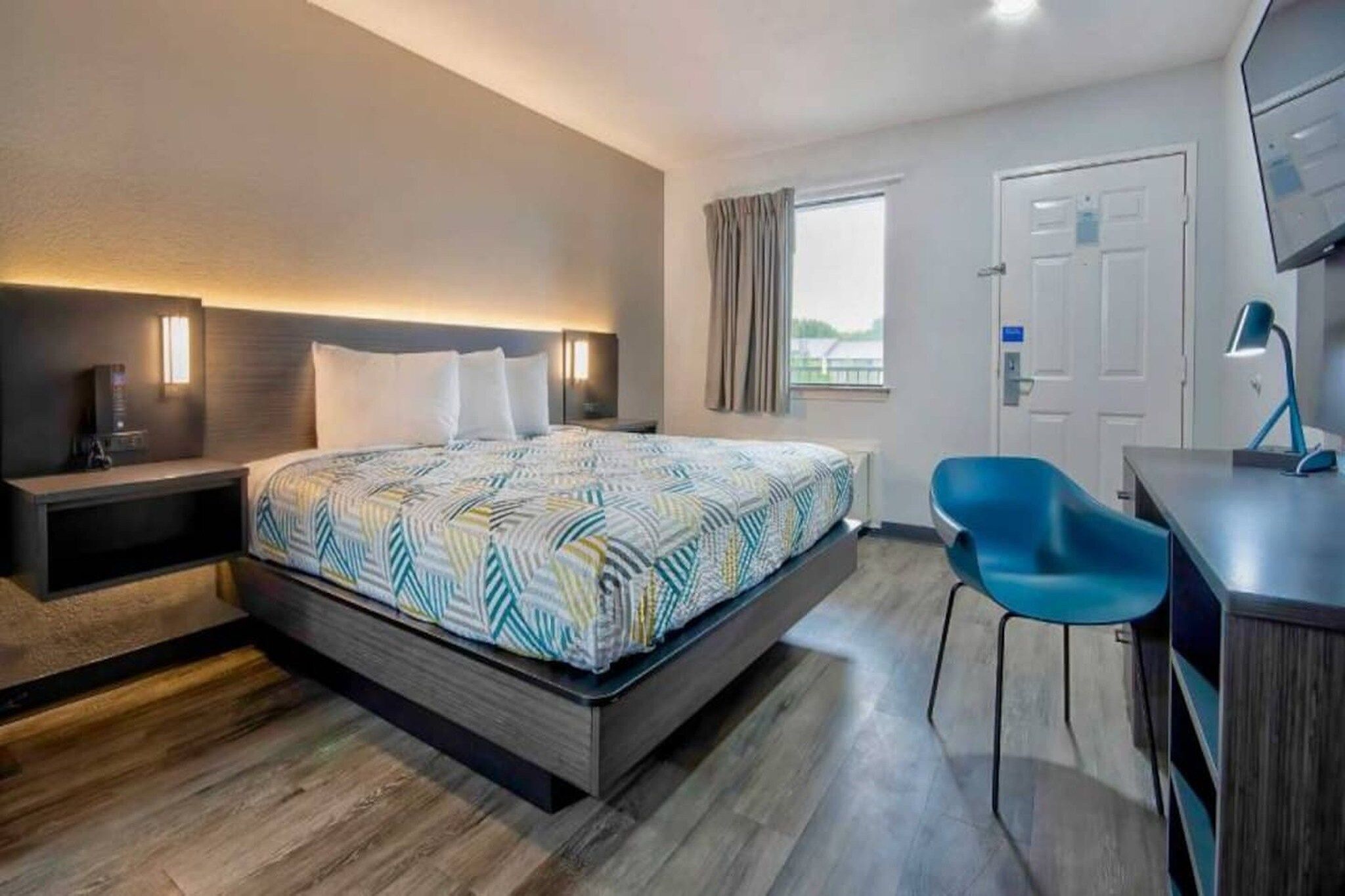 Une chambre d’hôtel comprenant un lit, une table de chevet, un bureau, une chaise et une fenêtre avec des rideaux.