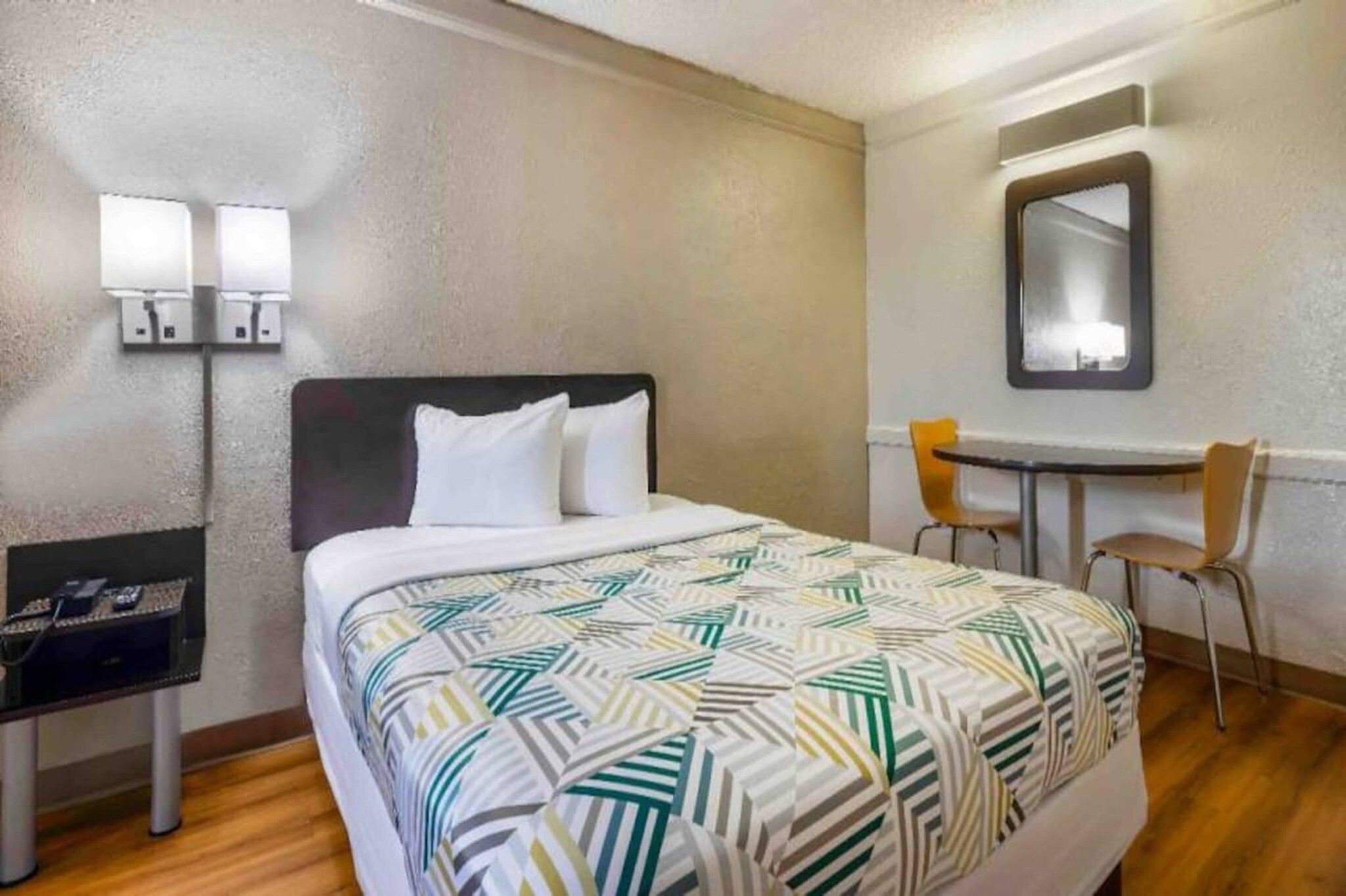 Una habitación de hotel con una cama, un escritorio, dos sillas, un espejo y una lámpara de pared.